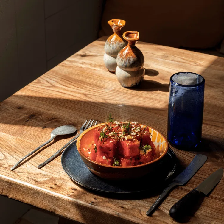 Wooden table with a bowl of cubed watermelon garnished with herbs and crumbled cheese, next to a blue glass and two ceramic pitchers, with a spoon, fork, knife, and butter knife placed around.
