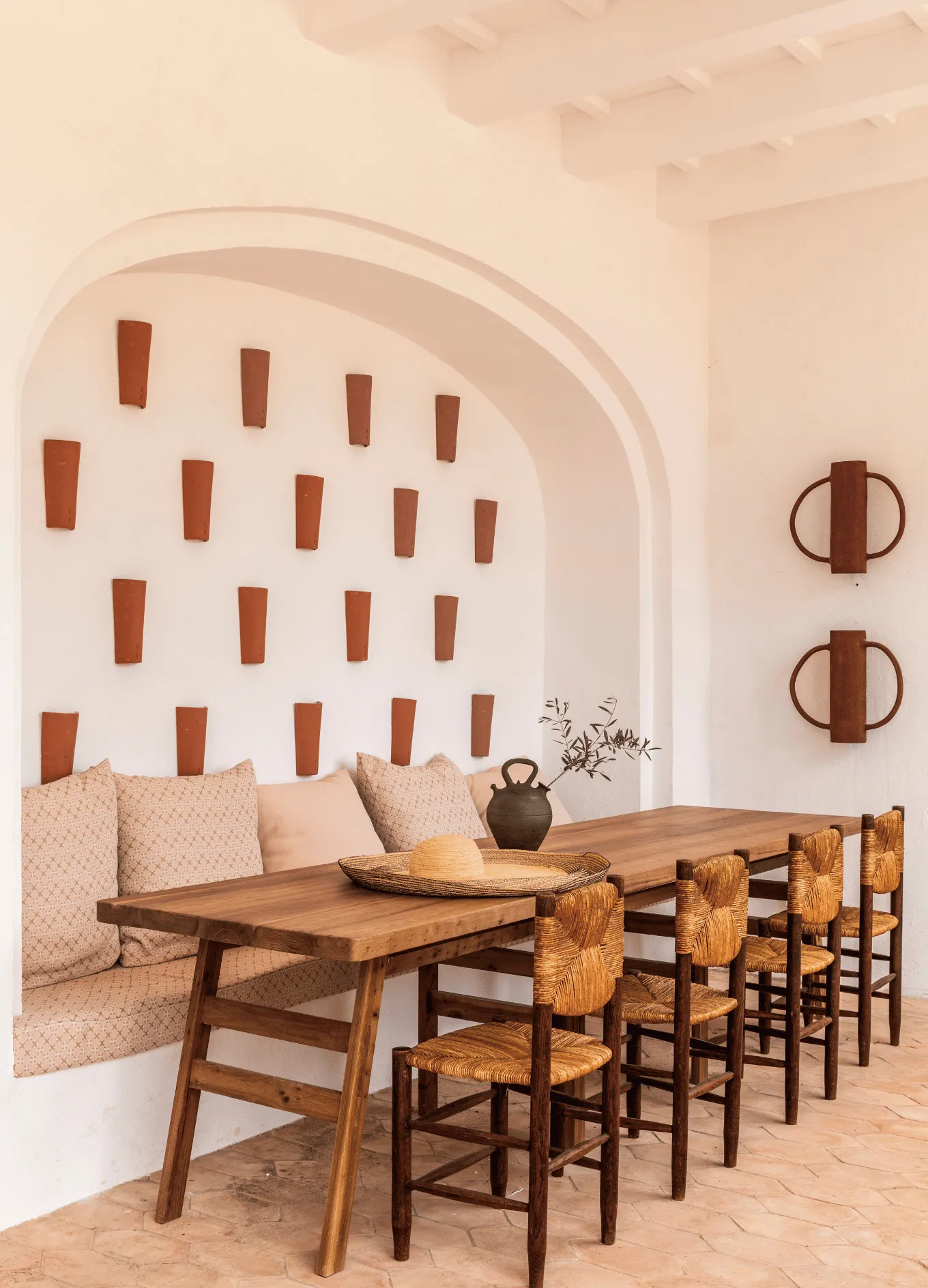 Wooden dining table with woven chairs and cushioned bench in a Mediterranean-style room with white walls and terracotta wall decor.