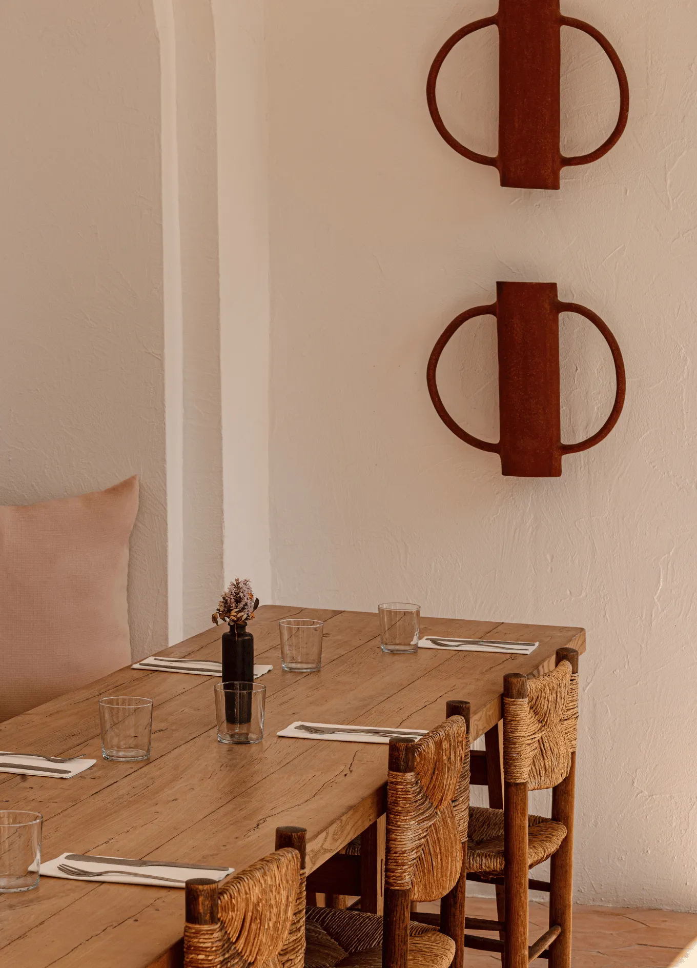 Wooden dining table set for four with glass tumblers and cutlery on napkins, surrounded by woven chairs against a beige wall with decorative circular metal wall hangings.