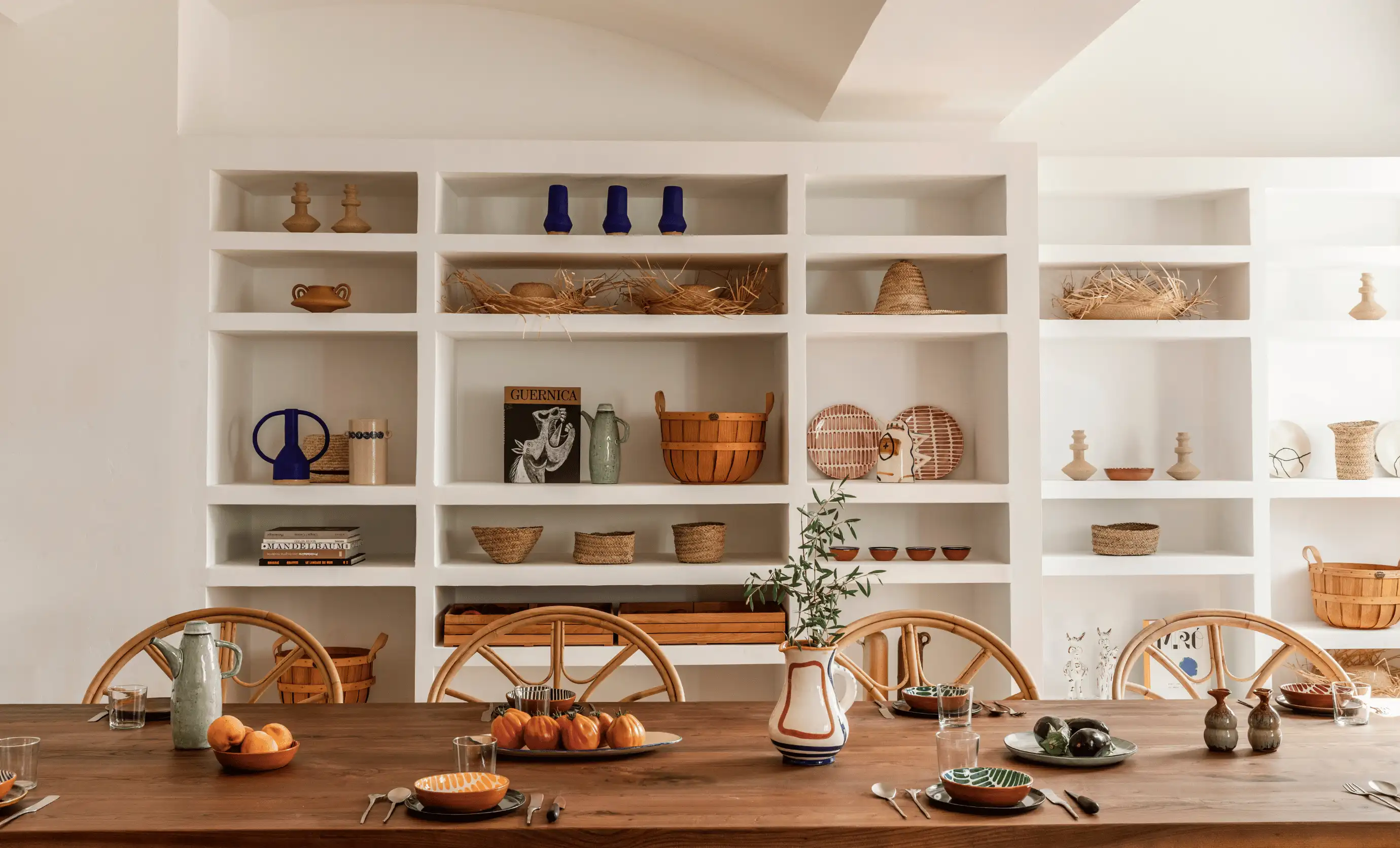 Dining table set with ceramic plates, glasses, and a vase with greenery in front of a white wall with built-in shelves displaying pottery, baskets, and decorative items.