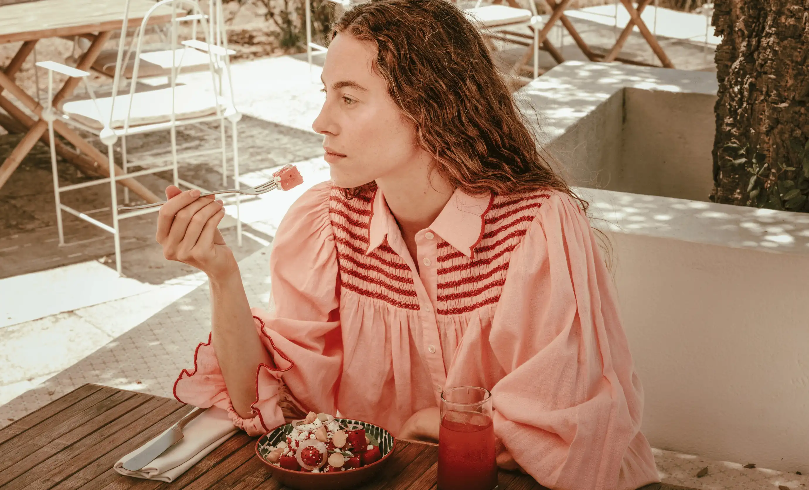 Woman in a pink blouse sitting at an outdoor wooden table, holding a fork with a piece of watermelon, with a bowl of watermelon salad and a glass of red drink in front of her.