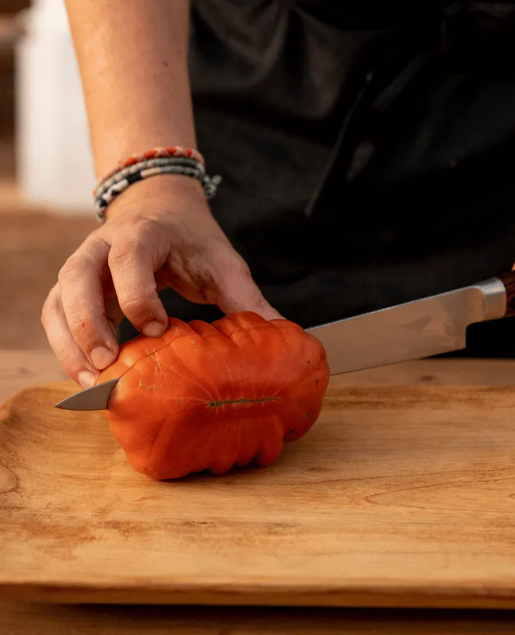 Hand holding a tomato with a knife inserted horizontally on a wooden cutting board.