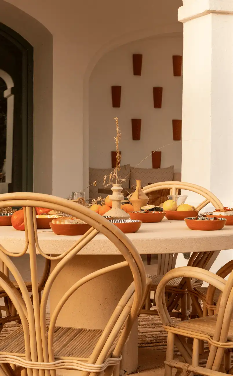 Outdoor dining table with beige stone top surrounded by rattan chairs, set with terracotta bowls of fruit and decorative vases in a Mediterranean-style space.