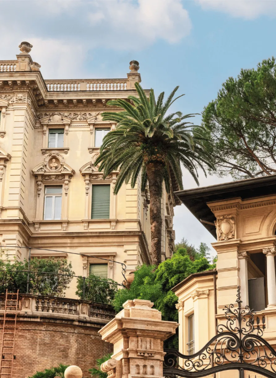 Ornate historic building facade with classical architectural details, a tall palm tree, and lush greenery under a partly cloudy sky.