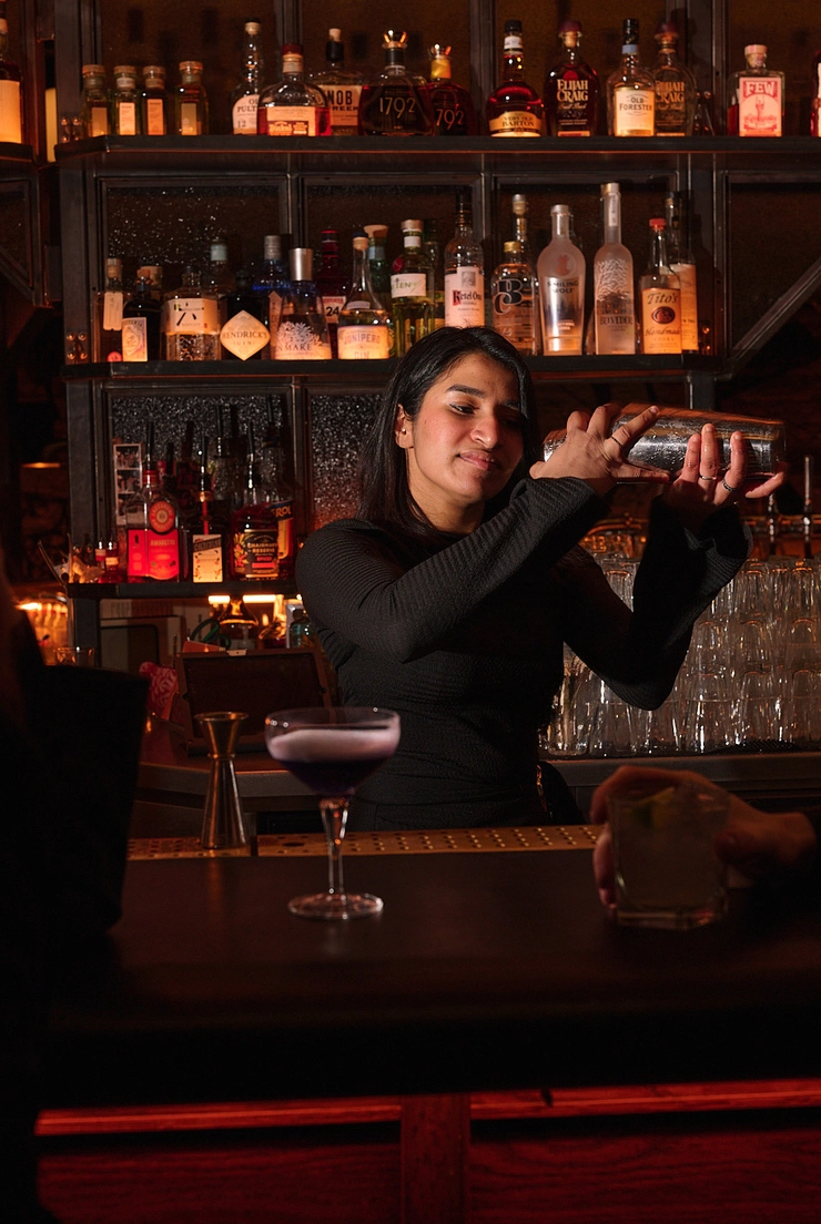 Bartender shaking a cocktail shaker behind a bar with shelves of liquor bottles in the background.