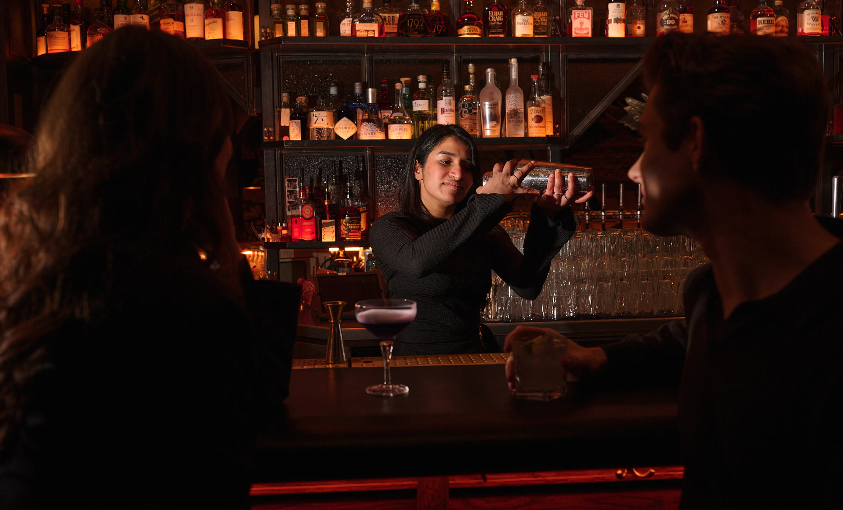 Bartender shaking a cocktail shaker behind a bar with two patrons watching, surrounded by bottles and glasses.