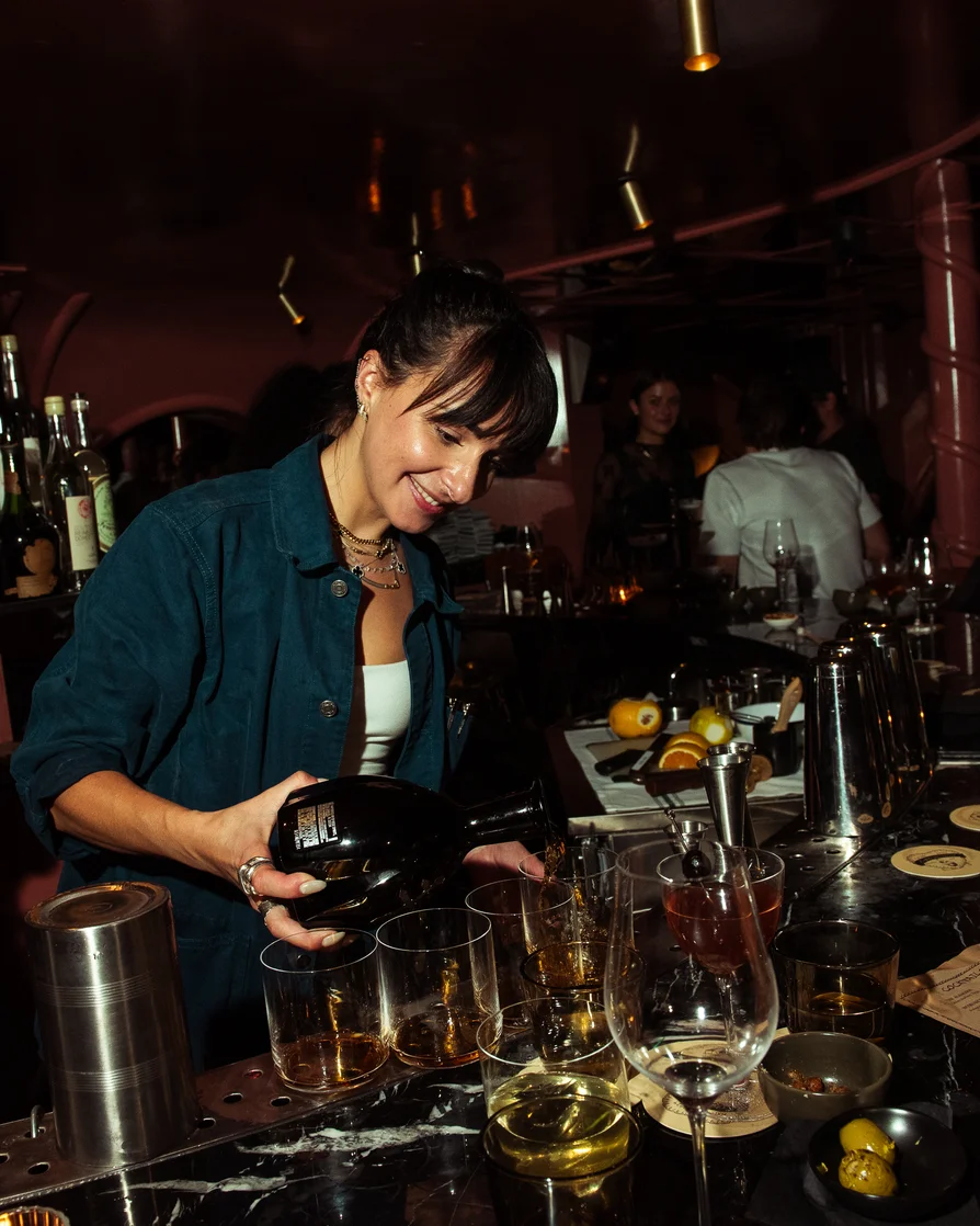 Bartender smiling while pouring drinks from a bottle into glasses at a dimly lit bar.