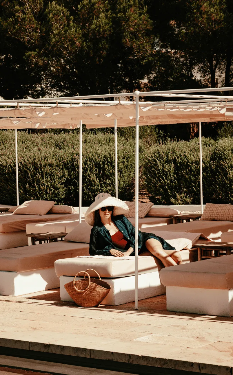 Woman wearing a large sunhat and sunglasses lounging on a cushioned outdoor daybed under a canopy near greenery.