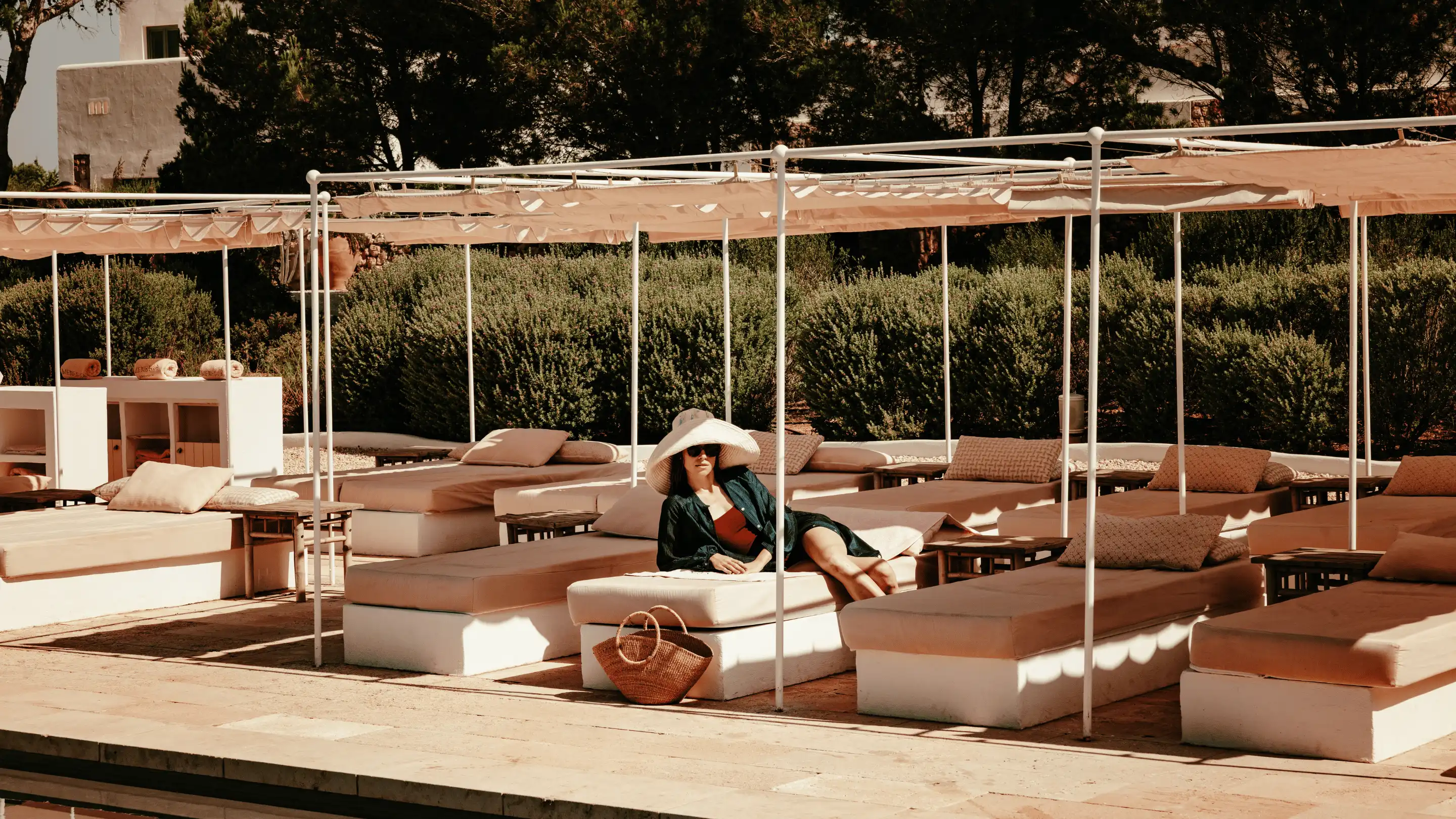 Woman in a large sunhat and sunglasses reclining on a poolside lounge chair under shaded canopies with cushioned seating and greenery in the background.