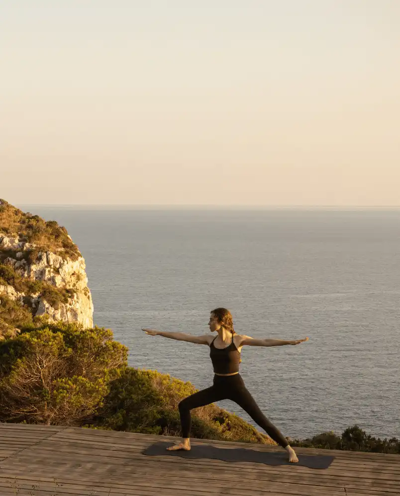 Woman in black workout clothes performing yoga Warrior II pose on a wooden deck overlooking the sea at sunset.