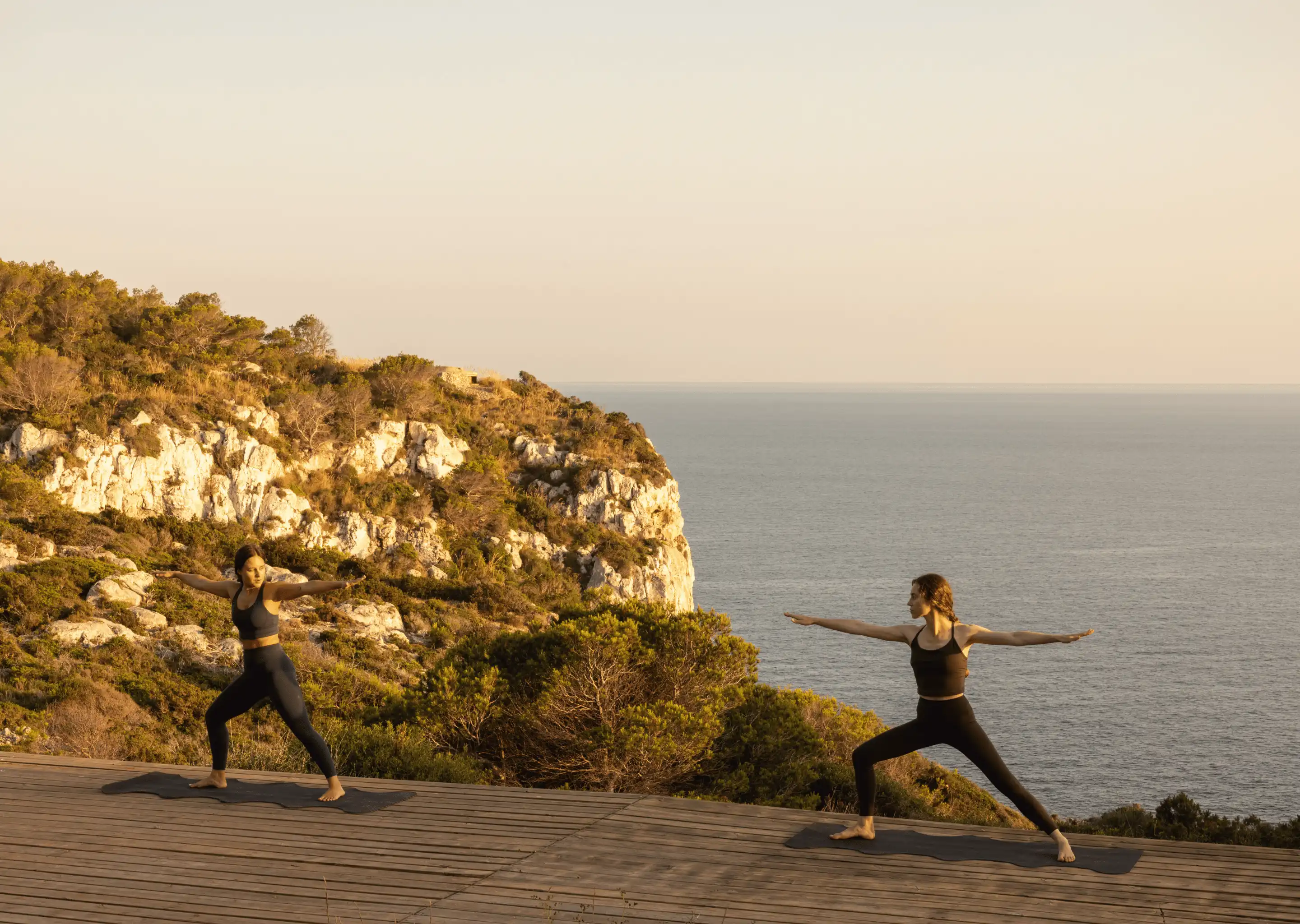 Two women in black workout clothes practicing yoga on mats on a wooden deck overlooking a rocky coastal cliff and calm sea at sunset.