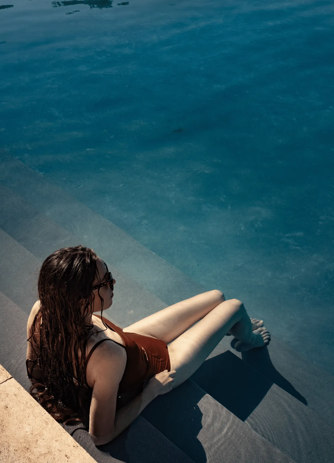 Woman in a brown swimsuit sitting on pool steps with wet hair and sunglasses, relaxing by blue water.