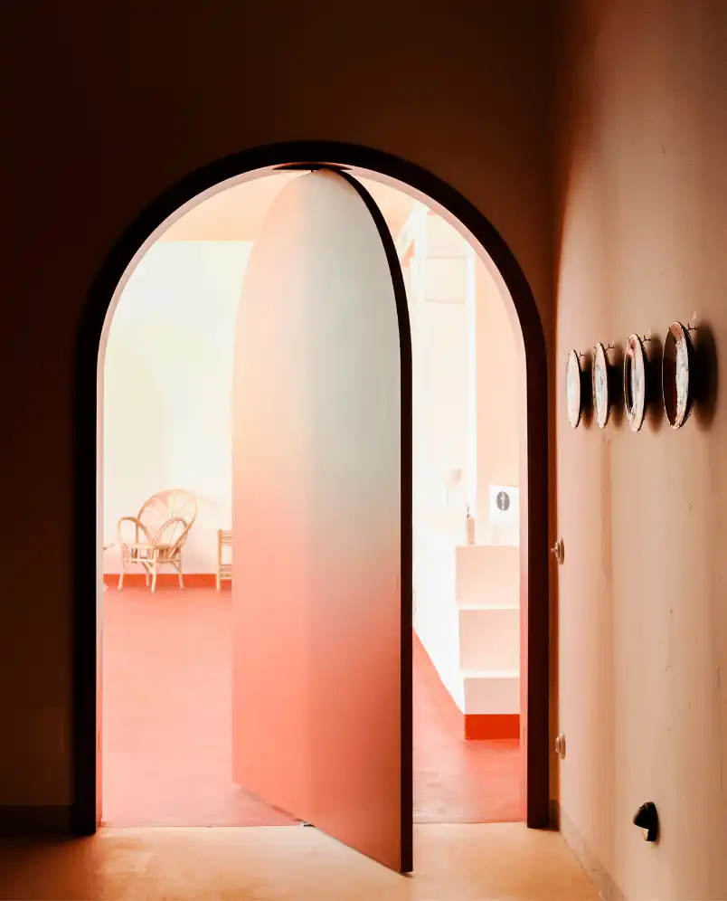 Partially open rounded door leading to a bright room with a wicker chair and terracotta floor.