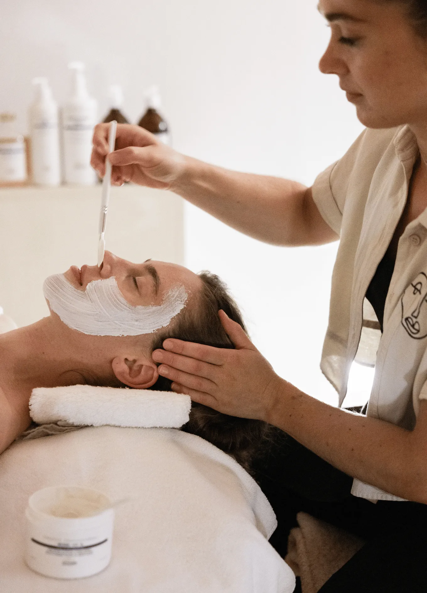 A woman receiving a facial treatment with a white cream being applied to her face by a spa therapist using a brush.