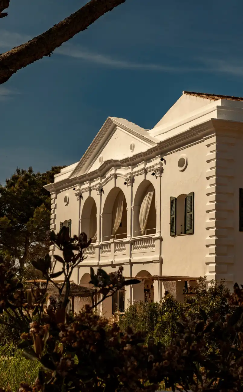 White Mediterranean-style building with arched balconies and green shutters surrounded by trees and plants under a clear blue sky.