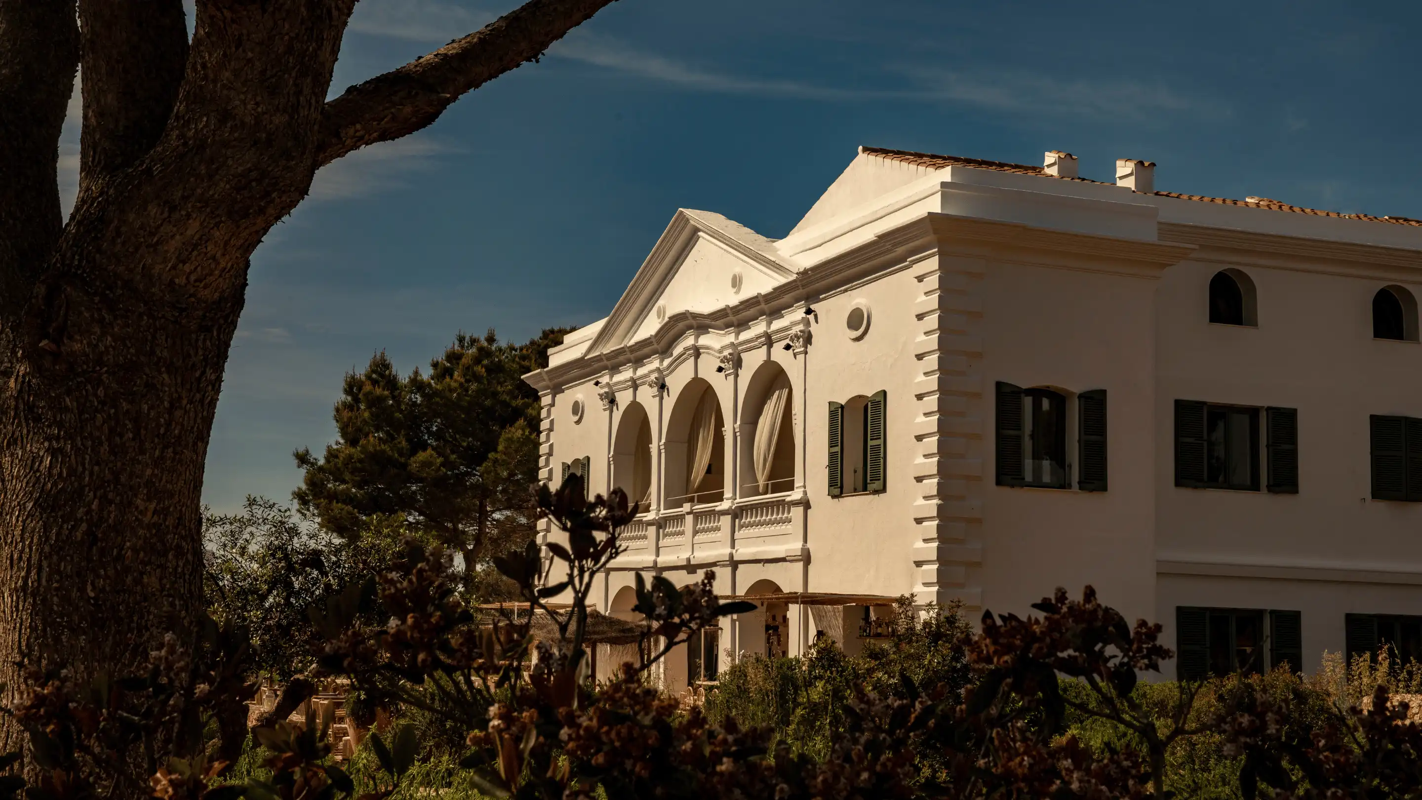 White two-story building with arched balconies and green shutters surrounded by trees and plants under a blue sky.