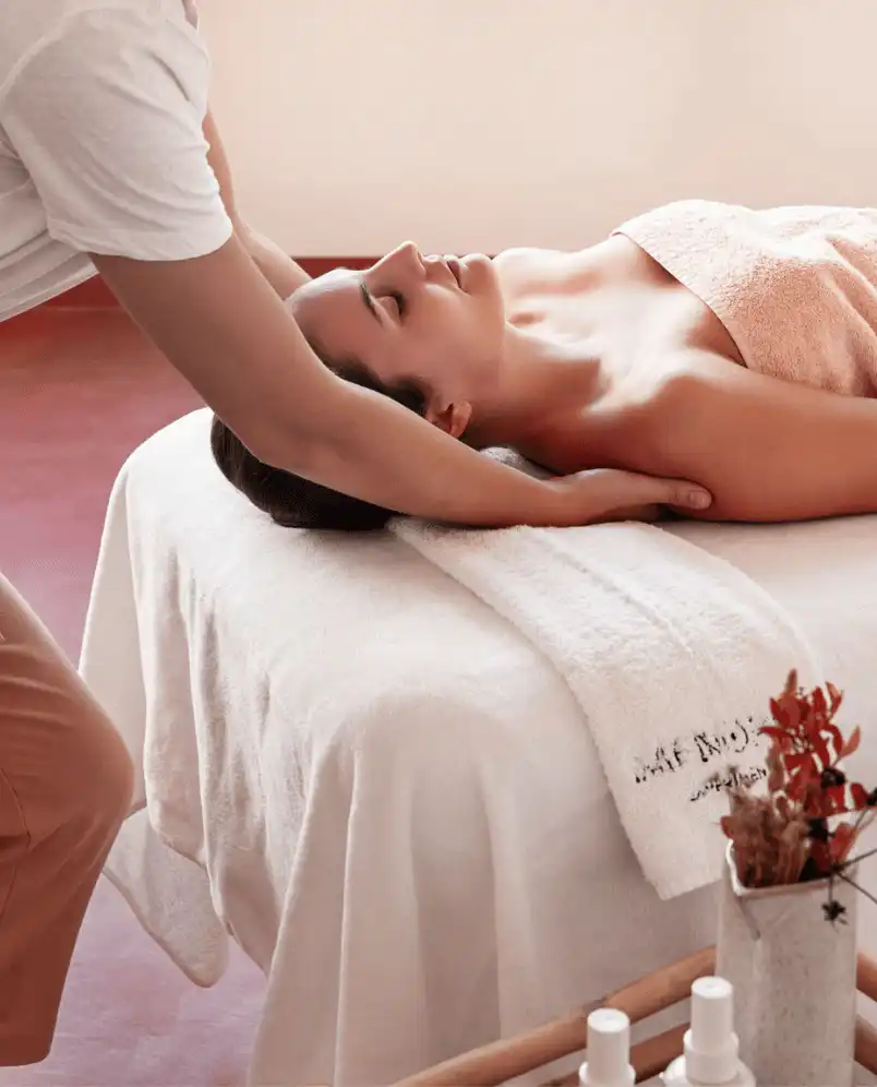 Woman lying on a massage table receiving a neck massage with a towel covering her chest in a spa setting.