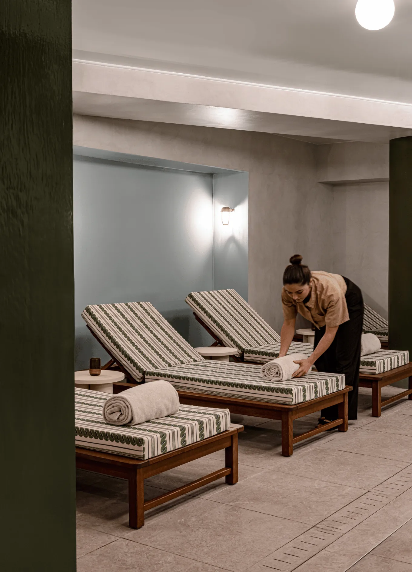 A woman arranging rolled towels on patterned lounge chairs in a calm spa relaxation area.