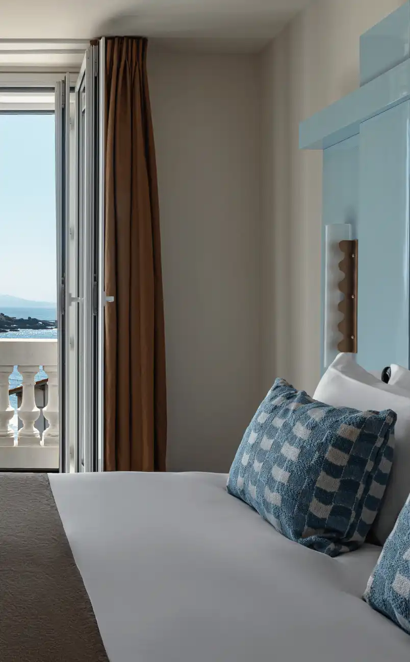 Hotel room bed with blue checkered pillows next to open window overlooking a balcony and ocean view.