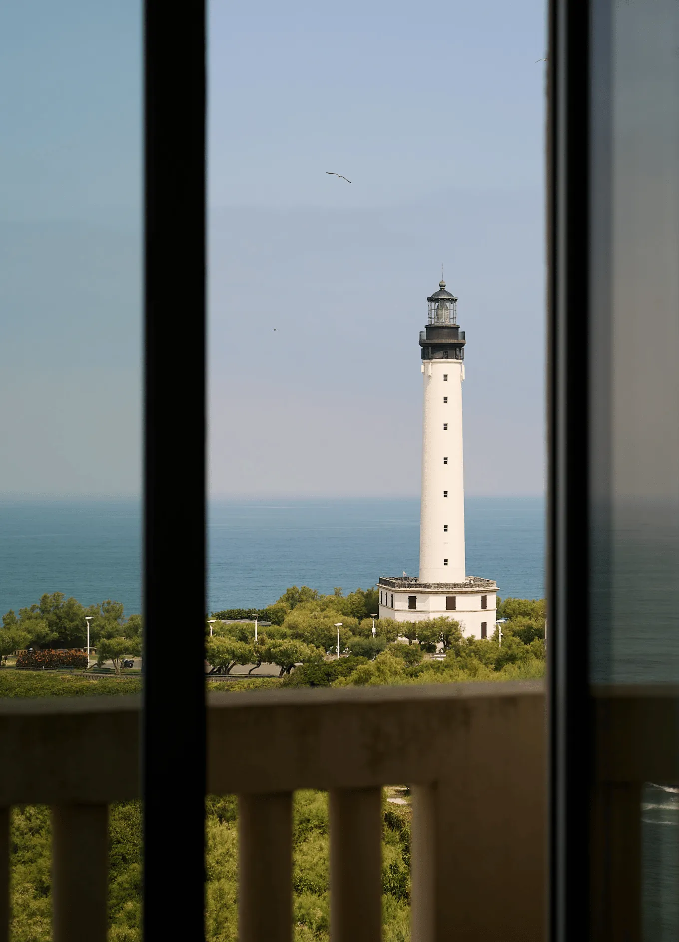 Tall white lighthouse with black top near coastline, viewed through a window with a balcony railing in foreground.