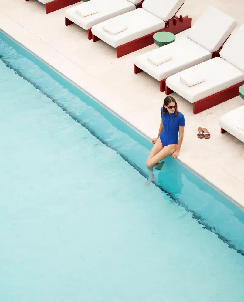 Woman in blue swimsuit sitting by the edge of a clear outdoor pool with white lounge chairs and small green tables nearby.