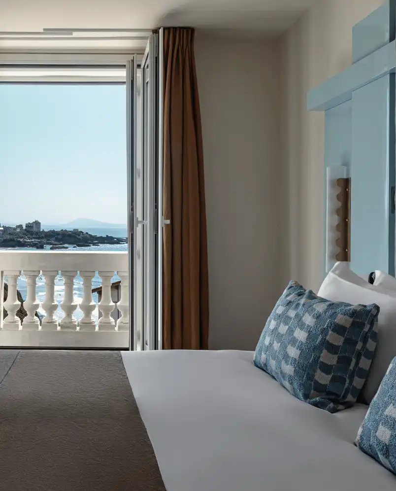 Hotel room bed with blue patterned pillows facing an open balcony door showing a sea view and distant coastline.