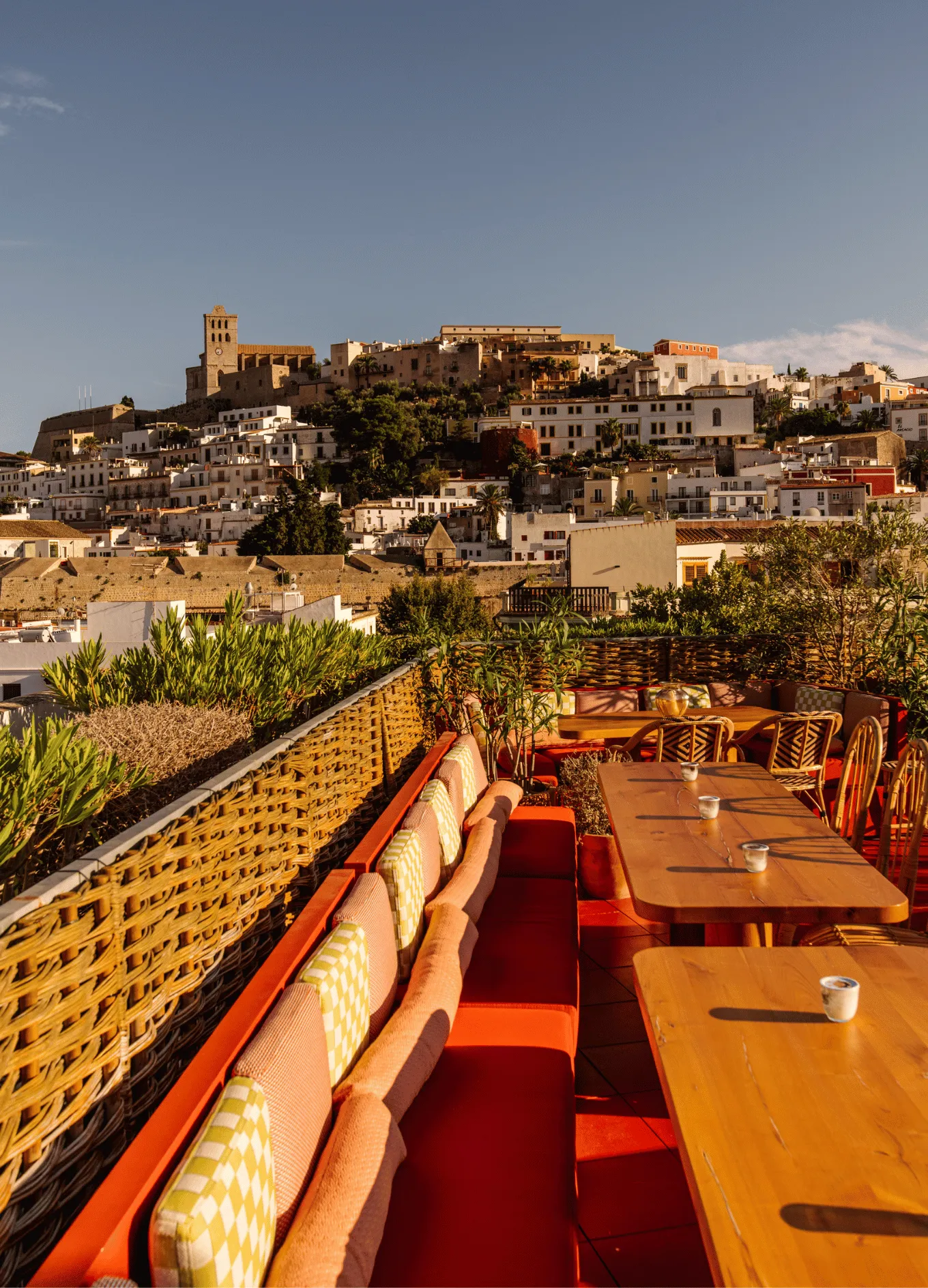 Terrace seating area with orange bench cushions and wooden tables overlooking a hill with white buildings and a historic stone church under a clear sky.