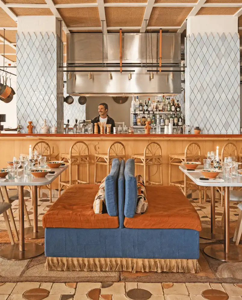 Modern restaurant bar with wooden stools, blue and orange cushioned seating, and a bartender standing behind the counter with shelves of bottles.