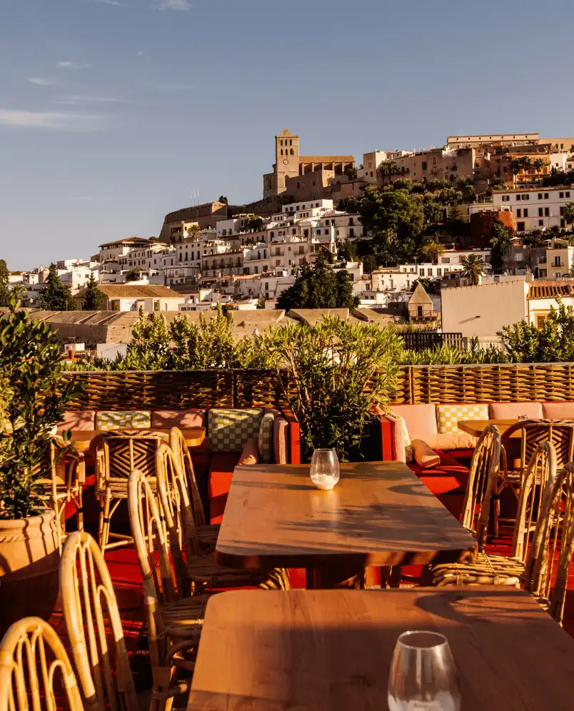 Outdoor seating area with wicker chairs and wooden tables overlooking a hillside town with a historic stone tower.