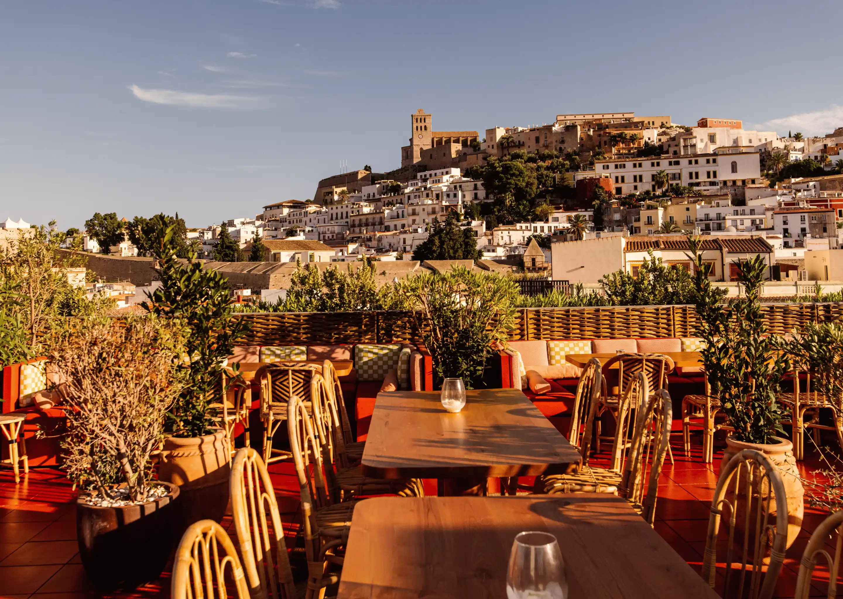 Outdoor terrace with wooden tables and wicker chairs overlooking a hillside town with white buildings and a castle.