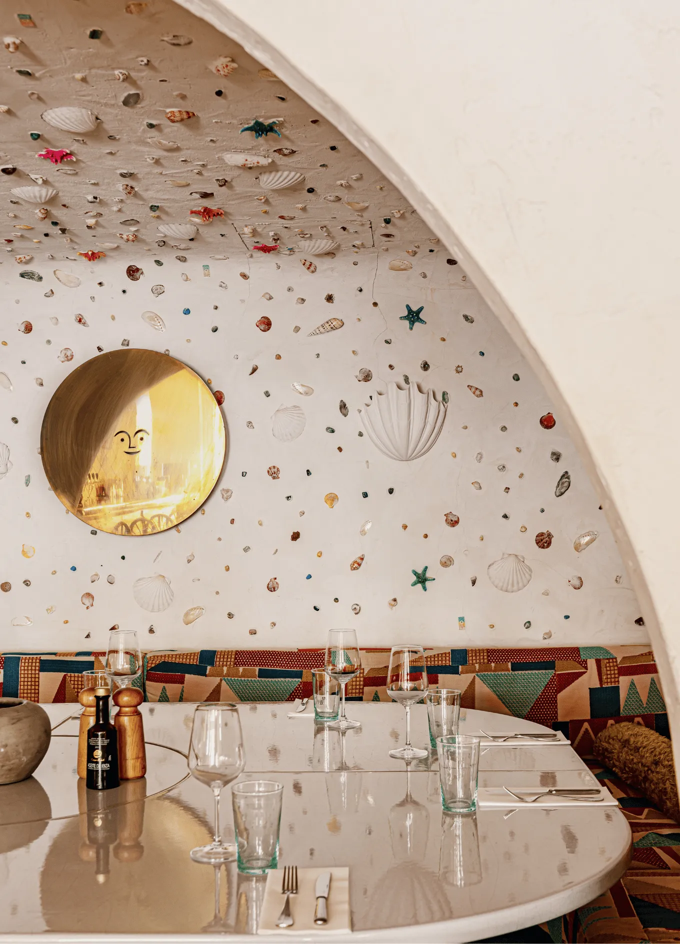 Curved dining table set with glassware and cutlery under an arched ceiling decorated with small seashells and starfish, with a round mirror on the wall.