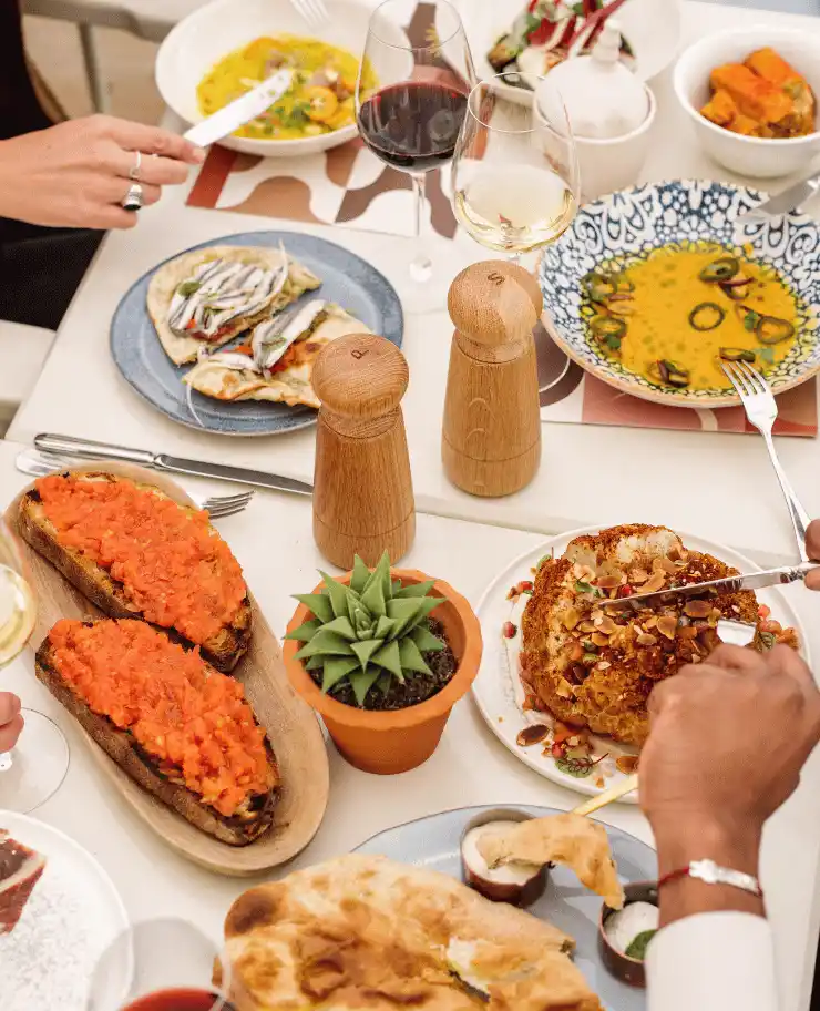Table set with a variety of dishes including toasted bread with red spread, grilled flatbreads topped with anchovies, a roasted cauliflower with nuts, a bowl of soup, and glasses of red and white wine.