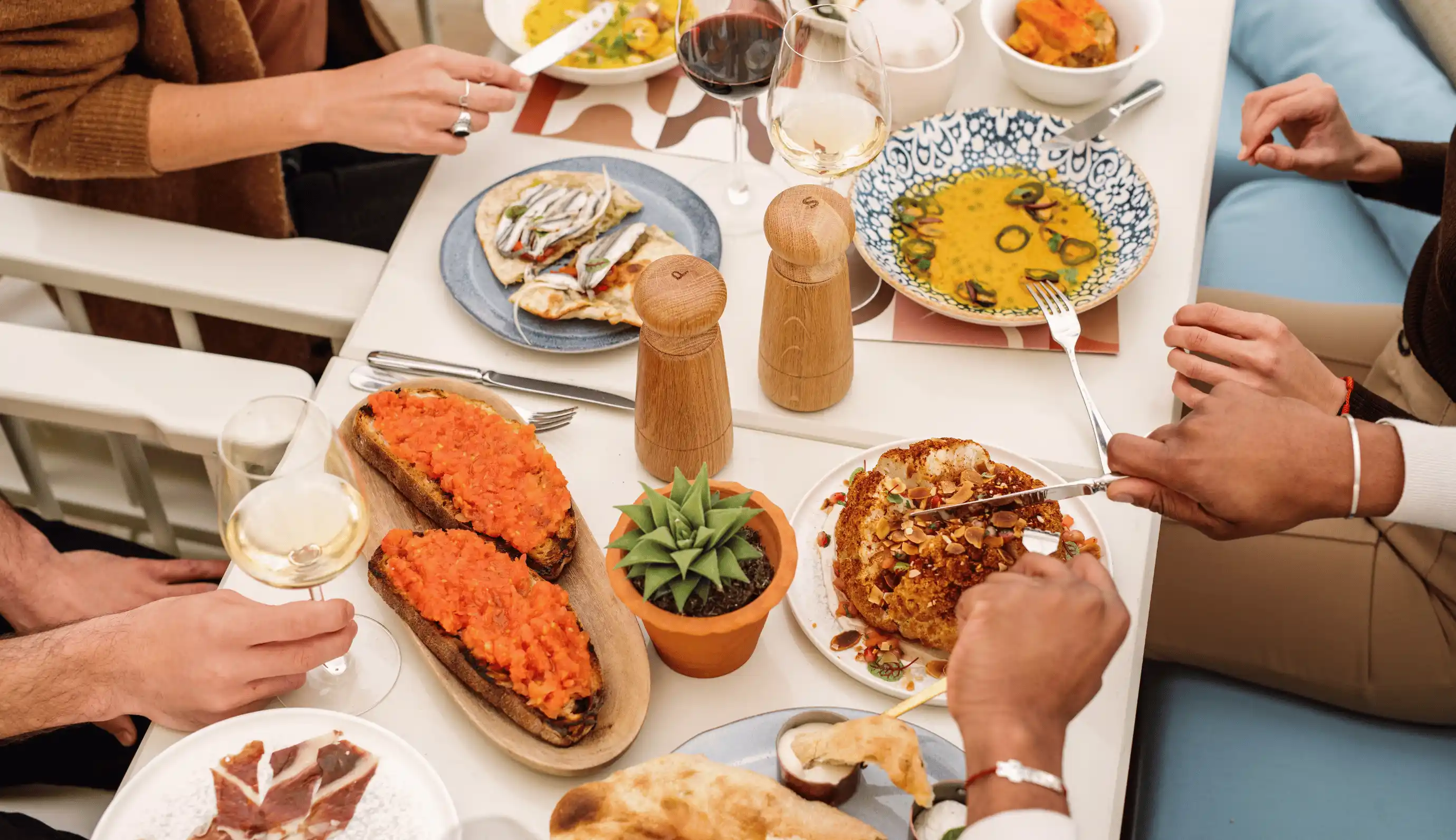People sharing a meal at a table with dishes including bruschetta, soup, roasted cauliflower, bread, and glasses of white and red wine.