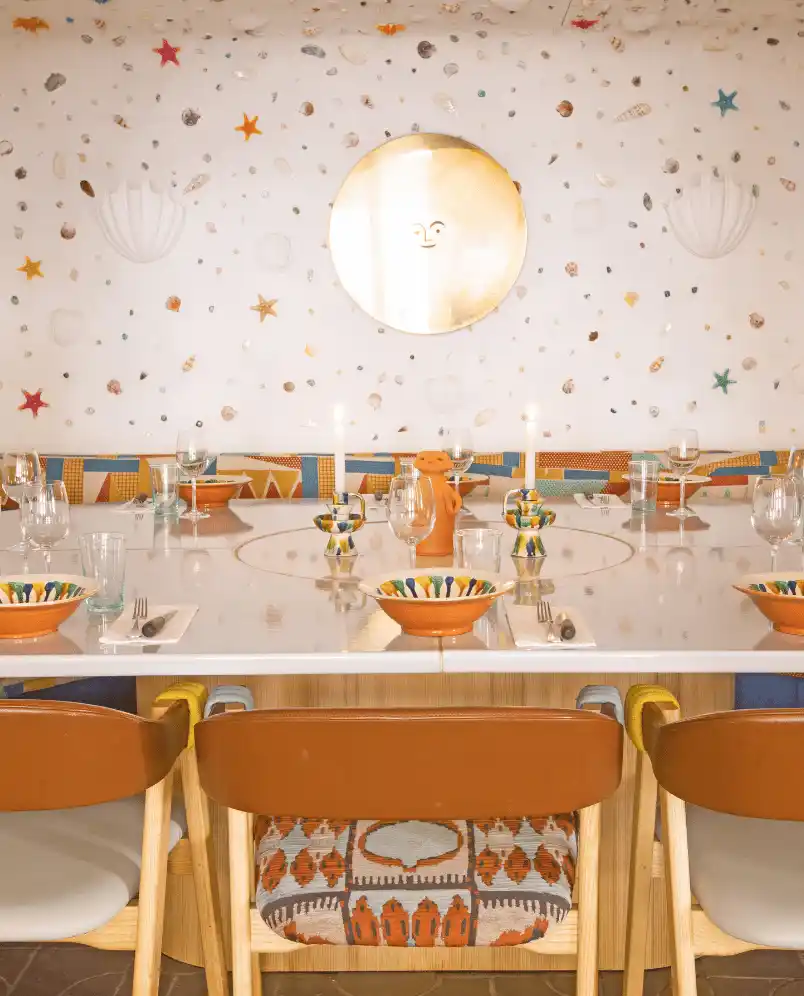 Cozy dining table set with colorful bowls, glasses, candles, and patterned chairs under a wall decorated with starfish and seashells around a sun face mirror.