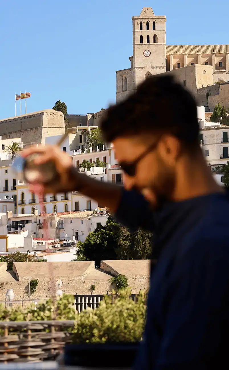 A man with sunglasses sprinkles reddish powder with a historic stone building and blue sky in the background.