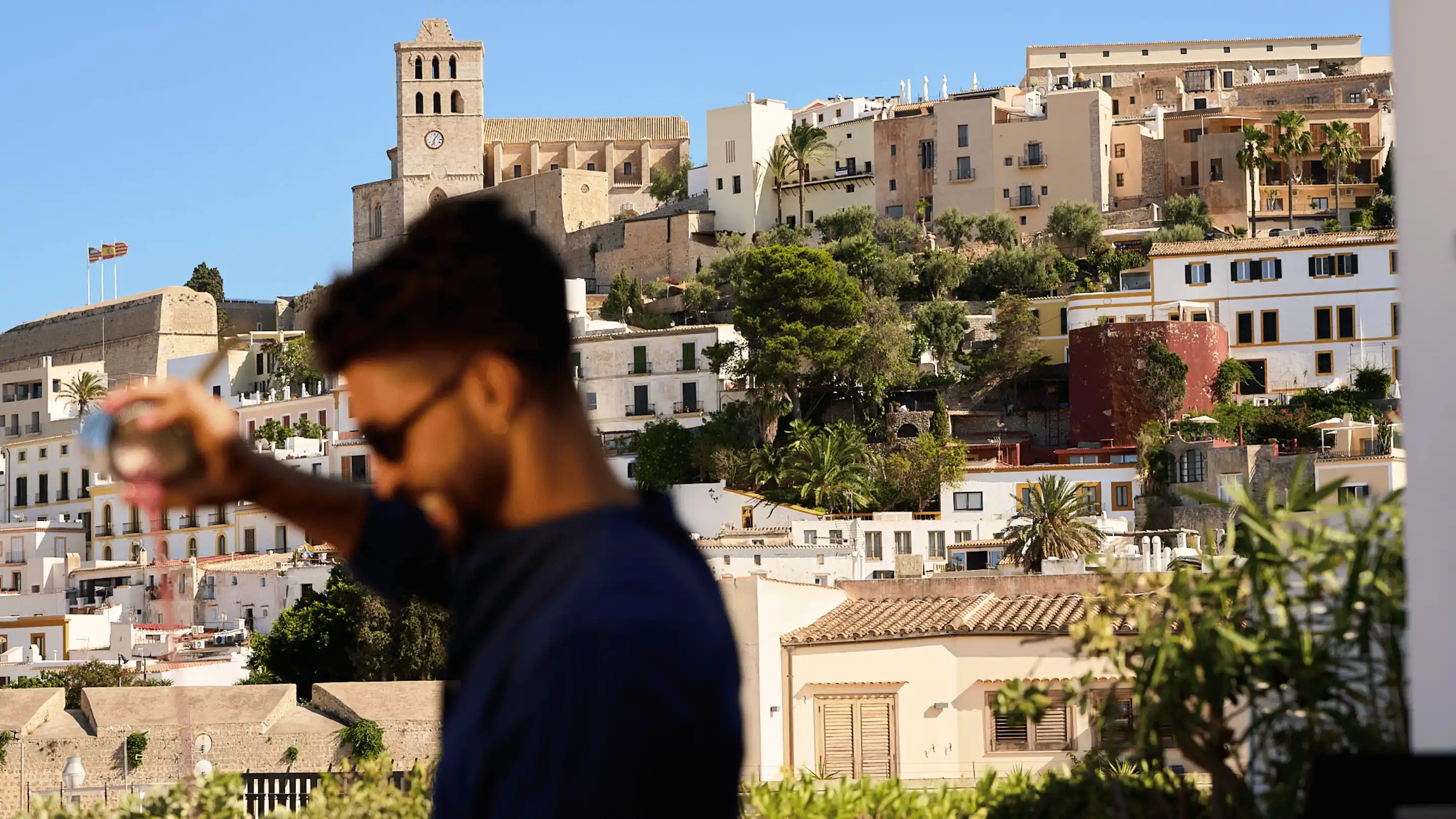 Blurred man wearing sunglasses holding a glass with a scenic view of Ibiza's historic buildings and a clock tower in the background.