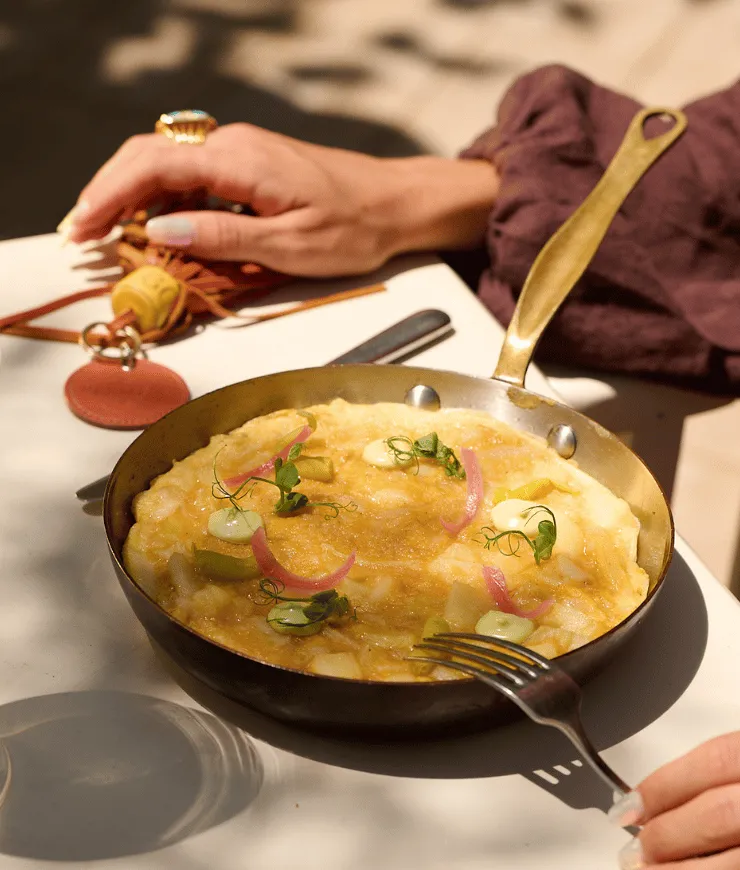 Frittata with green herbs and pink pickled onions in a brass skillet on a white table, with hands and a leather keychain nearby.