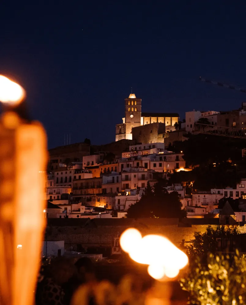 Night view of a hilltop castle and clustered white buildings illuminated by warm lights with blurred torch flames in the foreground.