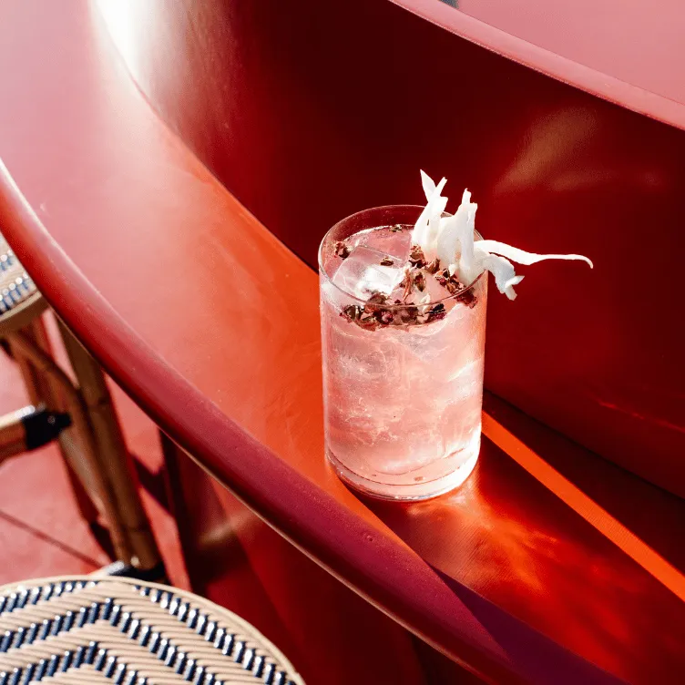 Clear cocktail with ice and white flower petals garnish on a curved red surface near a woven chair seat.