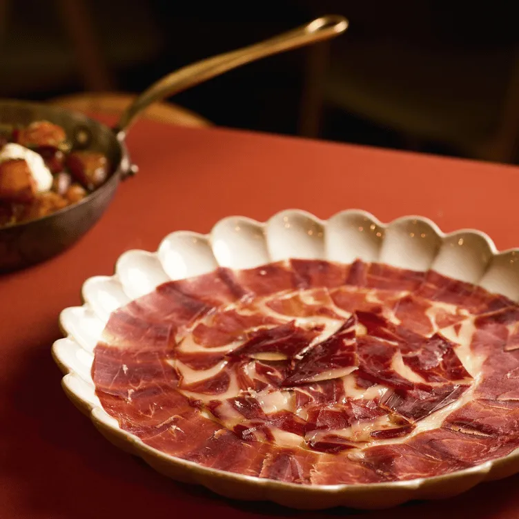 White scalloped plate with thinly sliced cured ham arranged in a circular pattern on a red tablecloth.