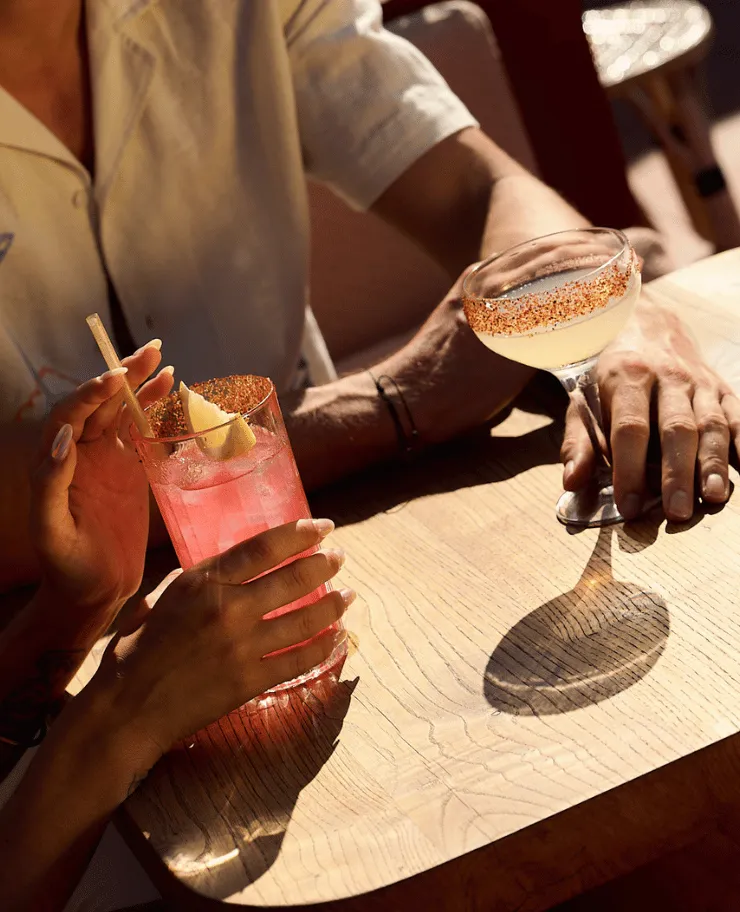 Two people holding cocktails at a wooden table, one drink pink with a lemon wedge and straw, the other a pale cocktail in a glass with a chili-salt rim.
