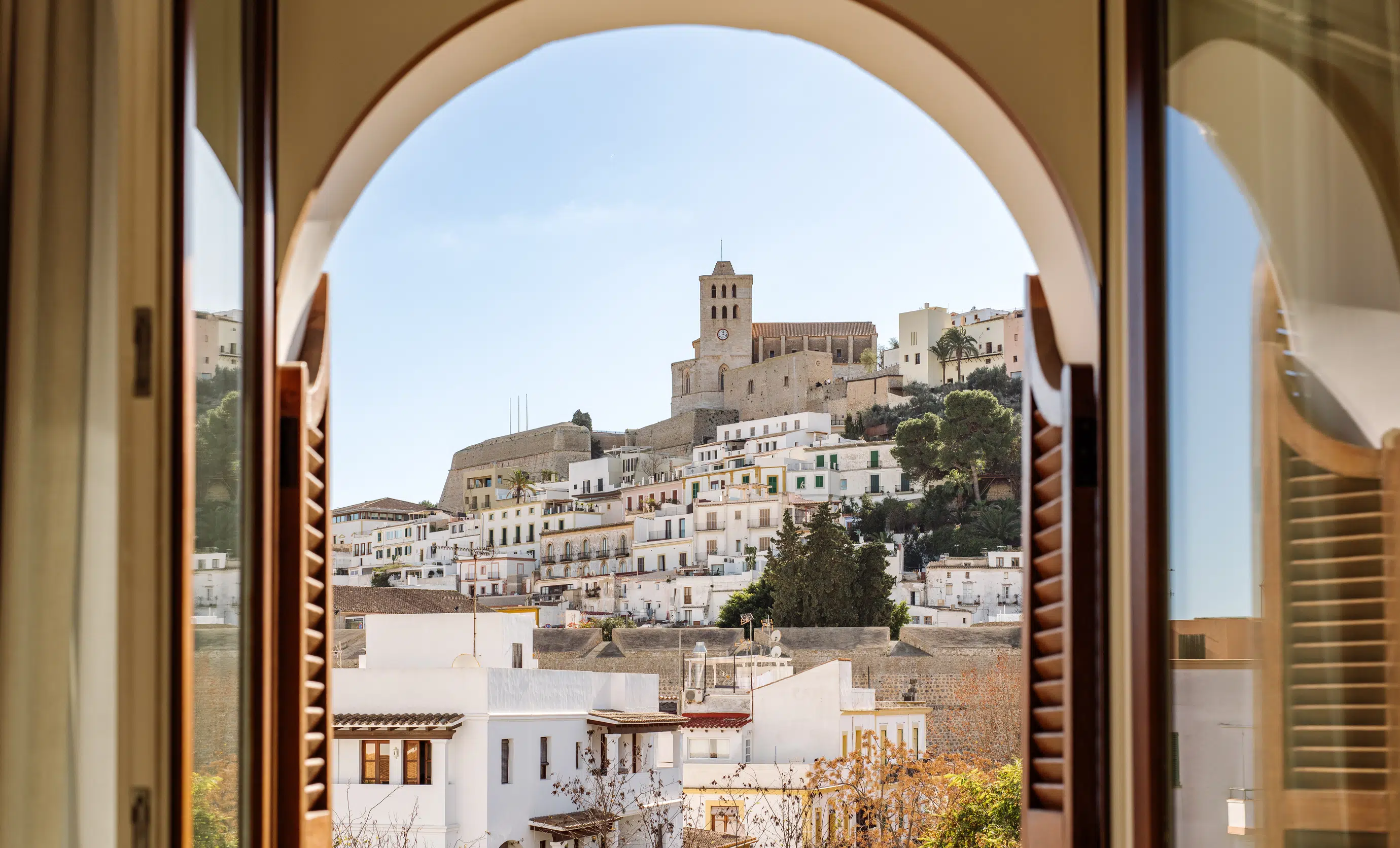 View of a historic hilltop church and white buildings framed by an arched window with wooden shutters.