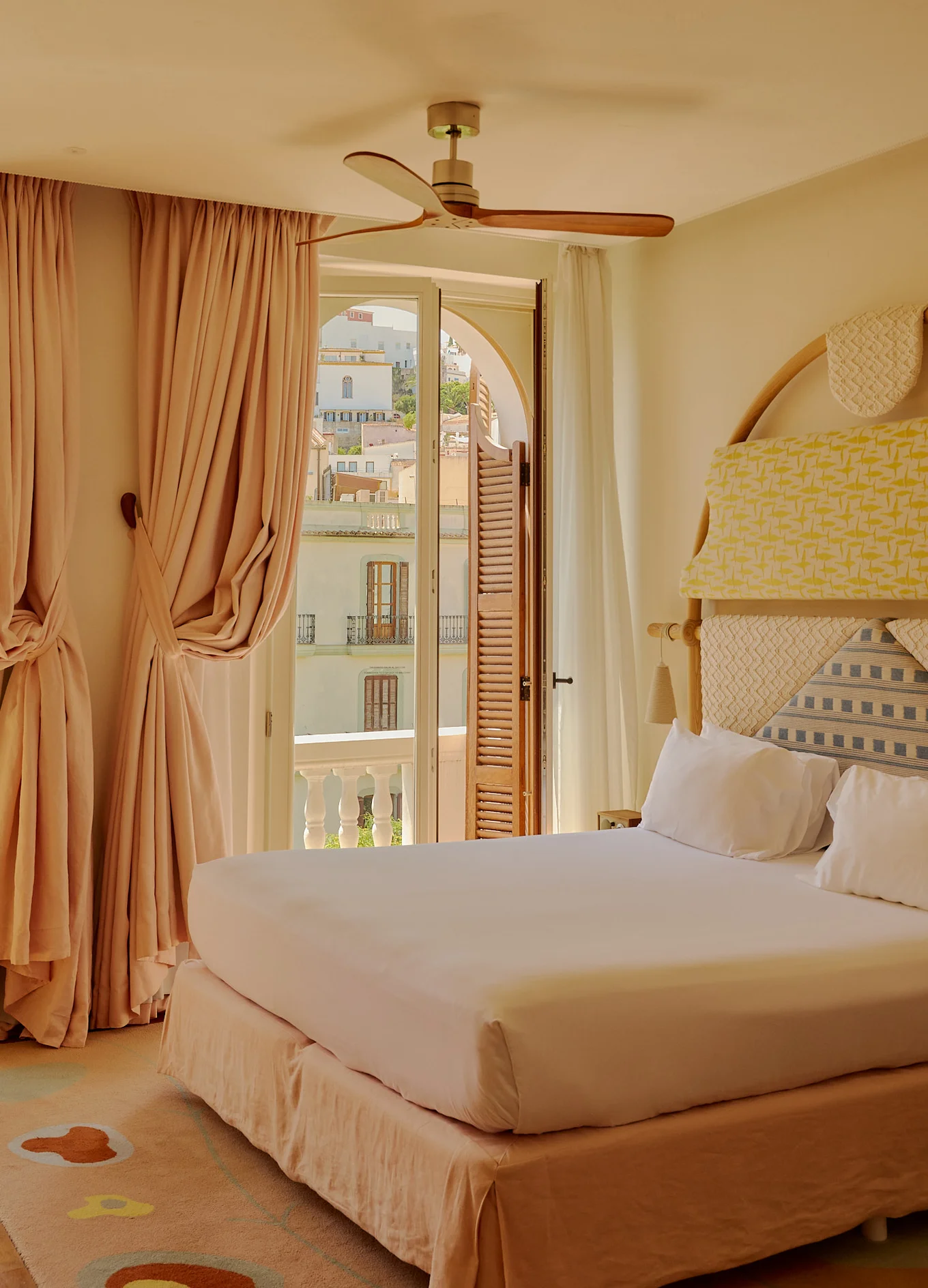 Cozy hotel bedroom with a large bed, colorful padded headboard, ceiling fan, and beige curtains beside balcony doors overlooking buildings.