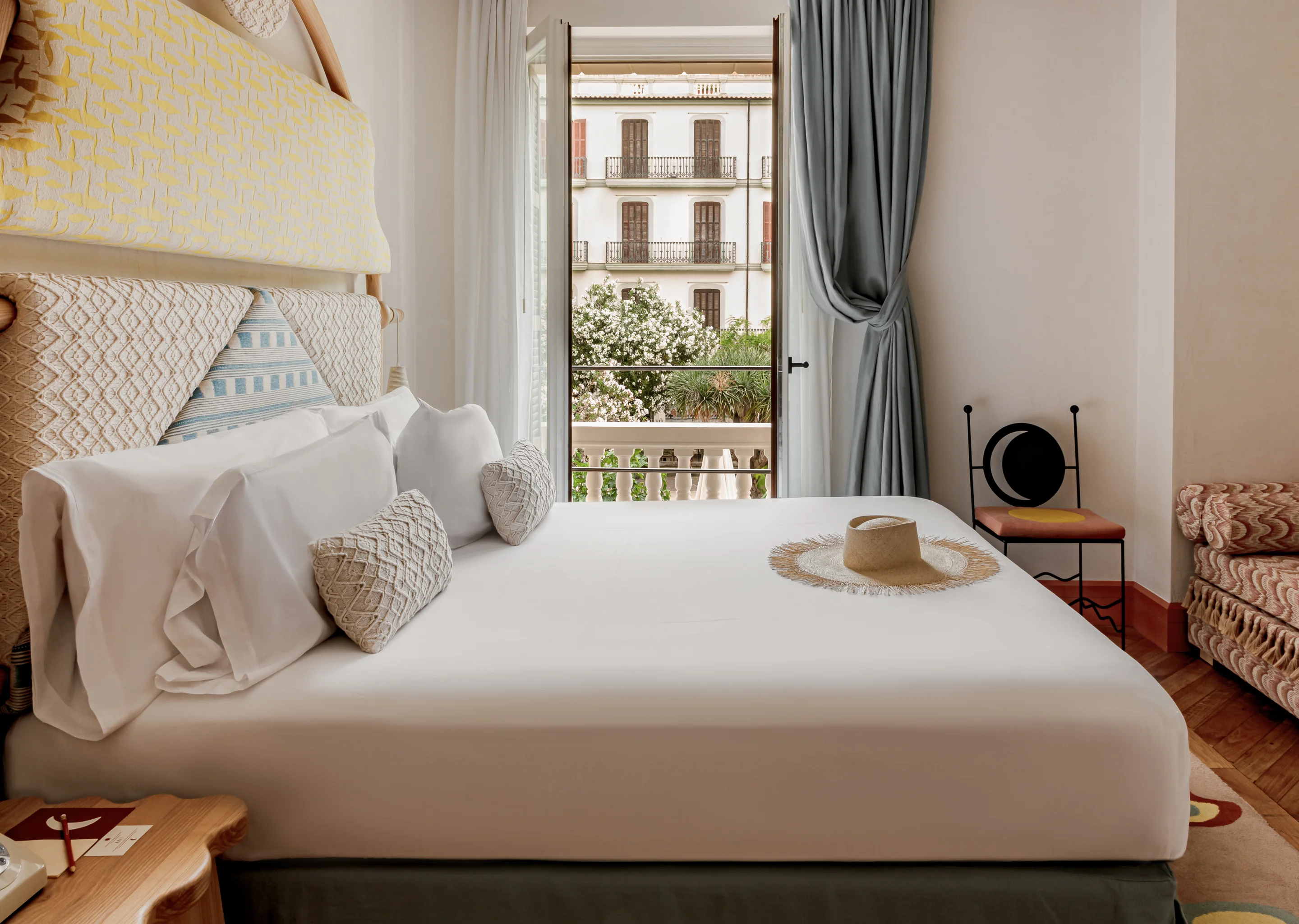 Bright hotel room with a large bed, decorative pillows, straw hat on white linen, open balcony door, and a chair near the window.