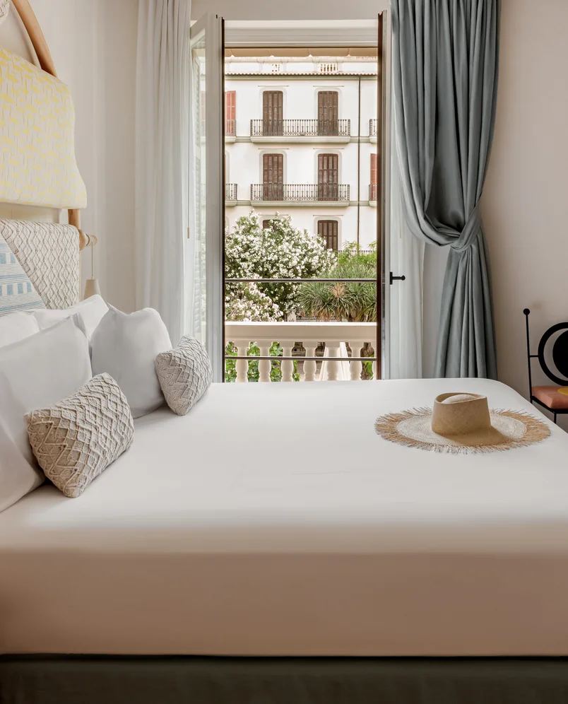 Bright hotel room with a neatly made bed, textured pillows, a straw hat on the bed, and open balcony doors revealing a building and greenery outside.