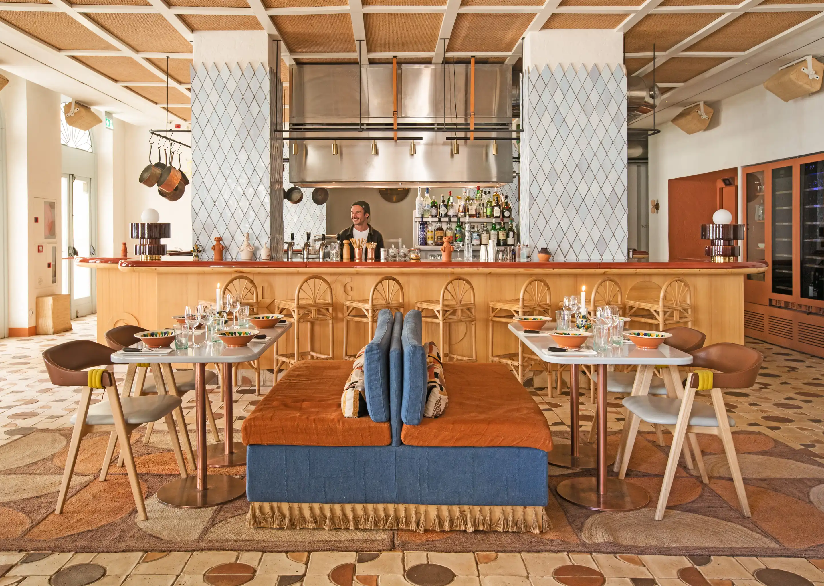 Cozy restaurant interior with a wooden bar, wicker bar stools, two tables set with bowls and glasses, and a smiling bartender behind the bar.