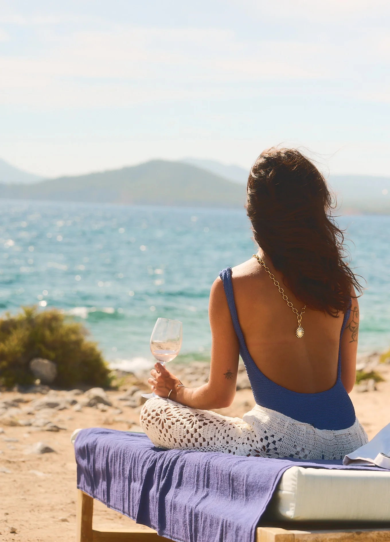 Woman in blue swimsuit and crochet pants sitting on a lounger by the beach, holding a wine glass and looking at the sea.