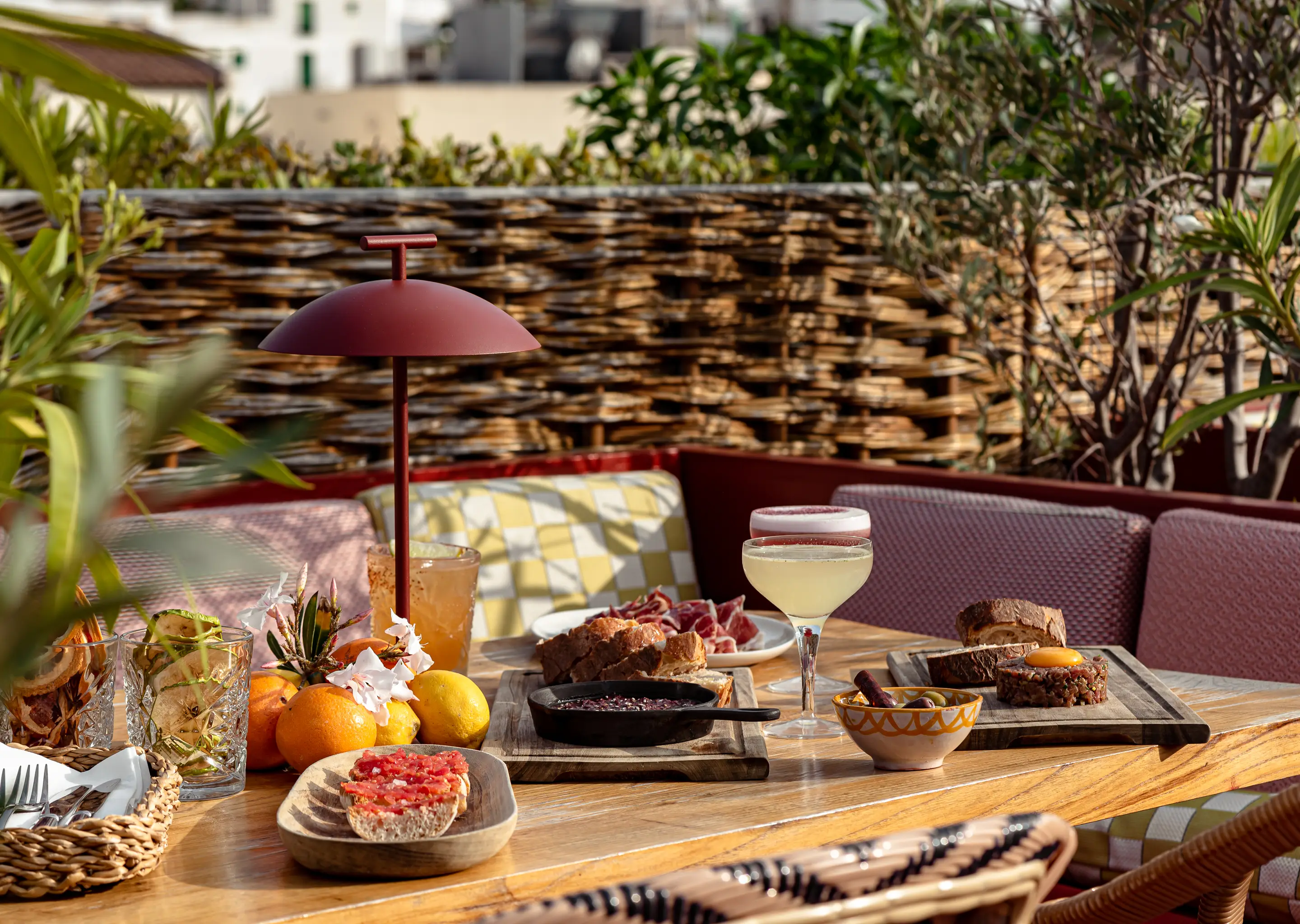 Outdoor wooden table with assorted appetizers including bread with tomato, sliced meats, steak tartare with egg yolk, olives, citrus fruits, and two cocktails in an inviting patio setting.