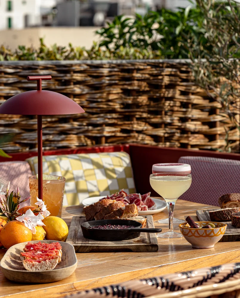 Outdoor table with assorted appetizers including bread with tomato, cured meat, a skillet dish, olives, citrus fruits, and two cocktails near patterned cushions and wicker fence.