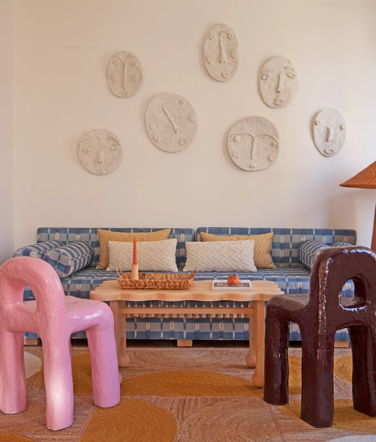 Living room with a blue checkered sofa, wooden coffee table, pink and brown sculptural chairs, and wall-mounted abstract face sculptures.